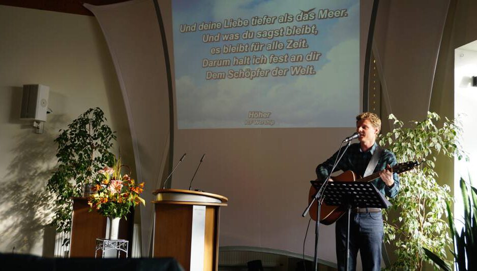 Mathias Feck steht mit Gitarre vorne am Mikrophon, im Hintergrund ein Liedtext