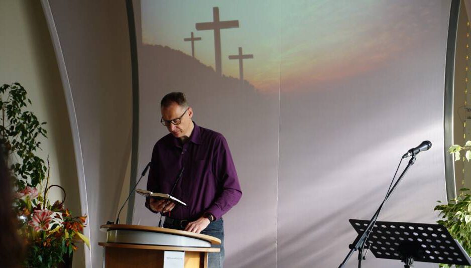 Joi Henn liest einen Text am Pult vor, im Hintergrund ein Bild mit drei Kreuzen