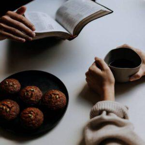 zwei Hände auf einem Tisch mit Muffins, eine Hand hält eine Bibel,, die andere eine Tasse